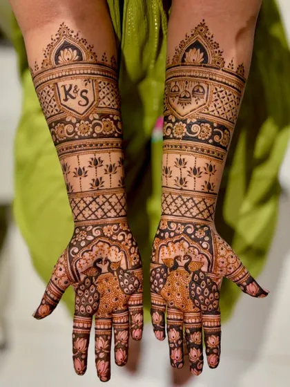 This intricate bridal design features the couple's initials, "K&S," inside a shield-like motif. The arms are adorned with lotus patterns and peacocks on the palms.