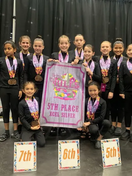 Way to go, Xcel Silver! This team secured 5th place at the Biles Invitational, celebrating with their medals and team banner.
