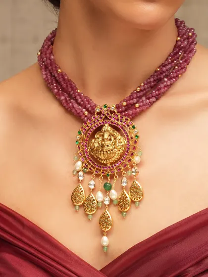 A divine temple jewelry piece, this necklace features a central deity motif surrounded by rubies and pearls, strung on multiple strands of ruby beads.