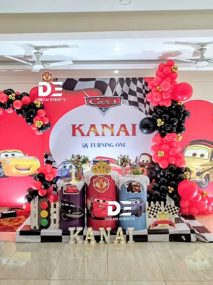 A full view of the Disney 'Cars' themed party for Kanai's first birthday. The setup features a racetrack backdrop, Lightning McQueen, and a red and black balloon arch with checkered flag details.
