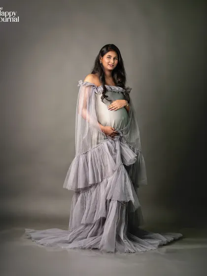 A full-length portrait against a solid grey background, making the texture and form of the gown the central focus. This is a classic, high-fashion maternity shot.