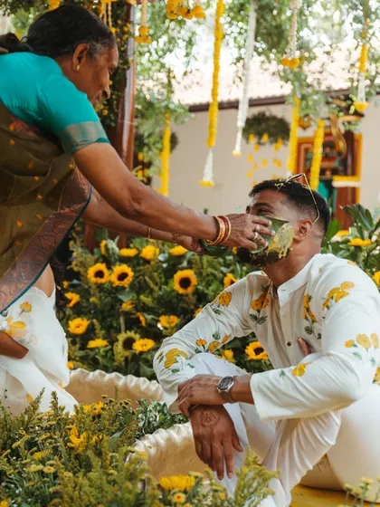 A tender moment where an elder applies turmeric paste to the groom. The background of bright yellow sunflowers adds to the warmth of the scene.