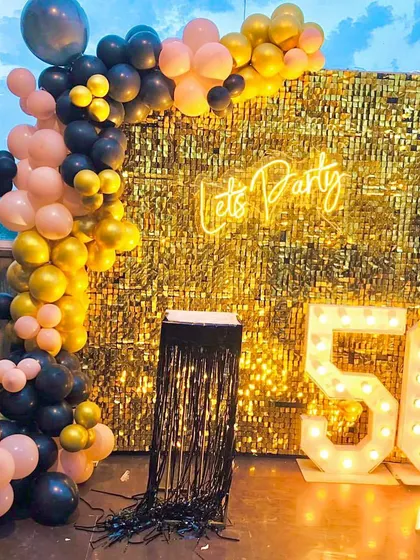 A 50th birthday party with a black, pink, and gold theme. The decor features a gold shimmer wall, a "Let's Party" neon sign, and marquee numbers.