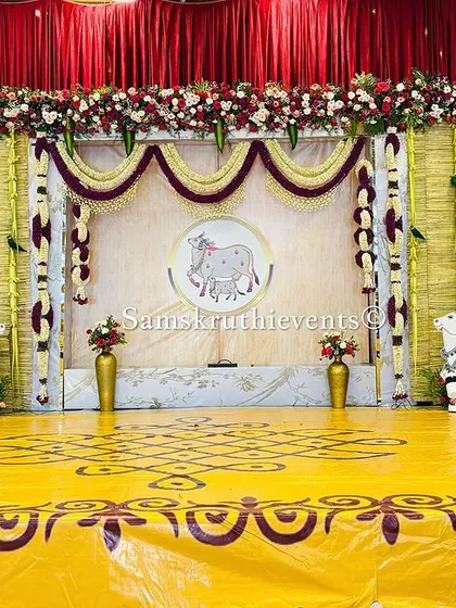 A grand stage setup featuring Pichwai-style paintings and cow figures. The backdrop is made of woven bamboo, and the stage is decorated with a traditional rangoli design, perfect for a large cultural event.