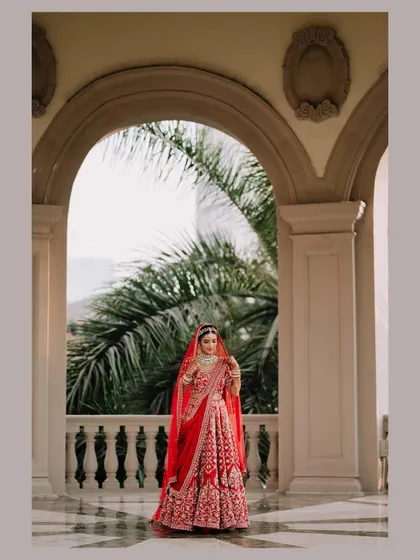 A stunning bridal portrait with the bride framed by a large archway, her red lehenga and long veil creating a striking silhouette.