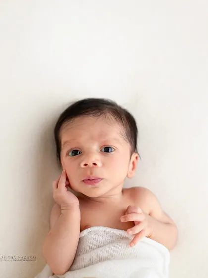 A two-week-old newborn looks up at the camera with a curious expression. The simple white wrap and background keep the focus entirely on the baby.