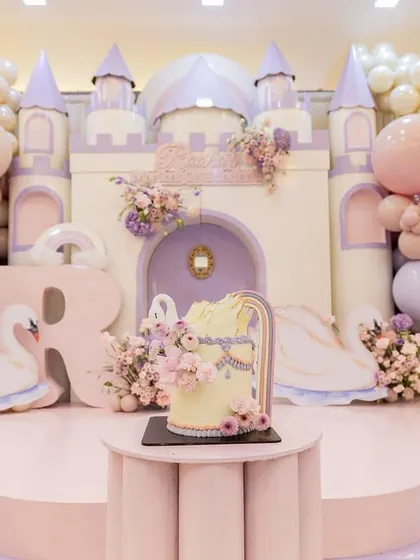 The main stage of the princess party, featuring a magnificent castle, whimsical swan figures, and a beautiful cake, all framed by pastel balloon clouds.