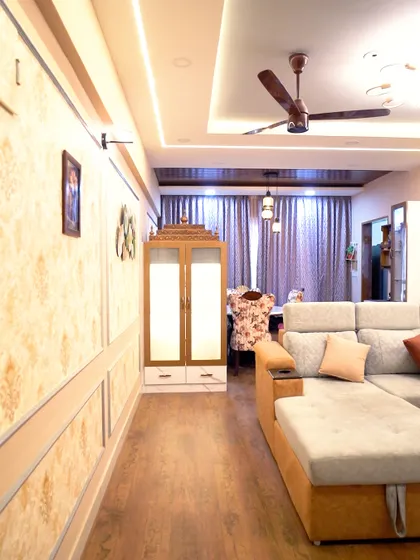 A luxurious living room with ornate wallpaper, a comfortable L-shaped sofa, and a dedicated pooja cabinet. The wooden flooring adds warmth to the elegant space.