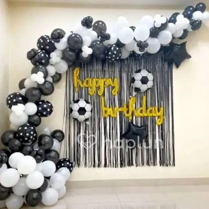 A football-themed birthday decoration. The black and white balloon arch is paired with football foil balloons and a "Happy Birthday" foil banner.
