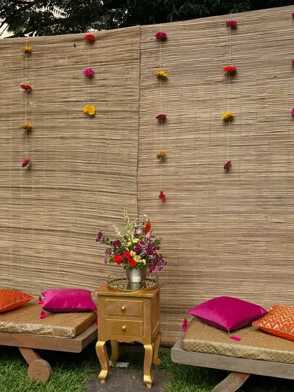 A simple and rustic seating arrangement for a Mehandi party. The low wooden benches with colorful cushions are set against a bamboo mat backdrop, creating a relaxed and traditional vibe.
