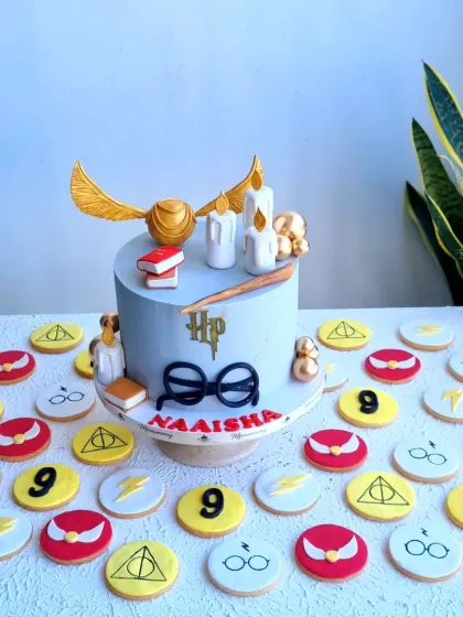 A magical Harry Potter themed birthday cake surrounded by custom-made cookies. The cake features iconic elements like the golden snitch, glasses, and a wand, while the cookies are decorated with symbols from the series, making it a perfect treat for any fan.