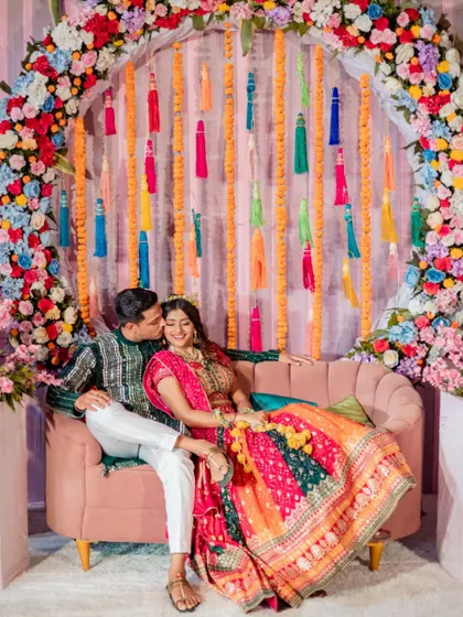 A sweet moment during the Mehendi ceremony, with the groom kissing the bride's cheek against a colorful backdrop.