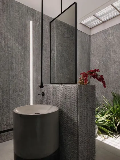 This powder room is a dramatic space, clad in grey stone with a freestanding basin. A skylight and a vertical light slit illuminate the textures, creating a moody and sophisticated atmosphere.