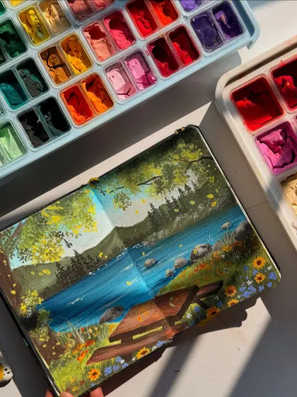 A top down view of my finished picnic scene in my gouache sketchbook. The sunlight on the page makes the colors of the wildflowers and the river look so bright.
