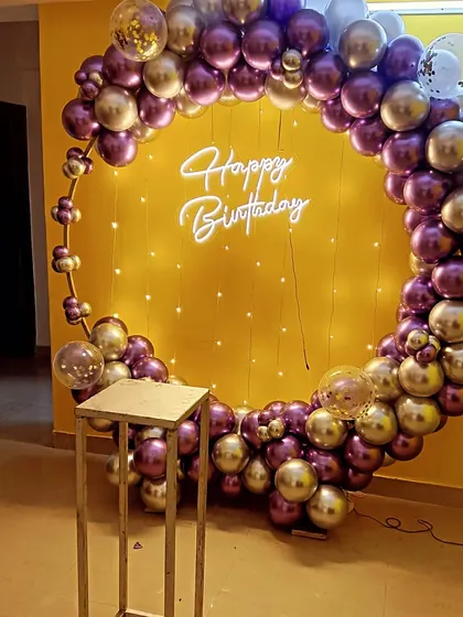 A purple and gold chrome balloon ring against a bright yellow wall, with a neon sign and fairy lights for a pop of color.