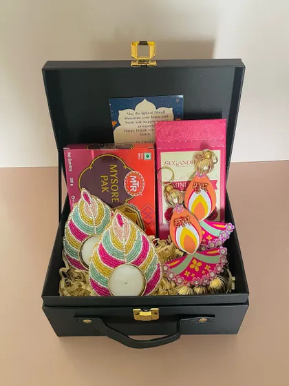 We love playing with colors. This black suitcase hamper features vibrant, beaded leaf-shaped t-light holders and traditional Mysore Pak, offering a unique blend of modern and classic.