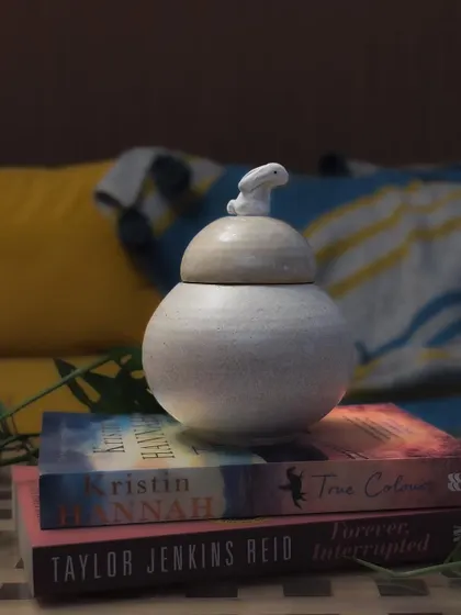 A serene, speckled white jar with a tiny rabbit on the lid, resting on a stack of books. This piece combines functionality with a subtle, charming design.