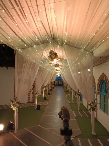 A beautiful walkway decorated with white drapes and fairy lights. The path is lined with floral arrangements on pedestals, guiding guests towards the main event area with elegance.