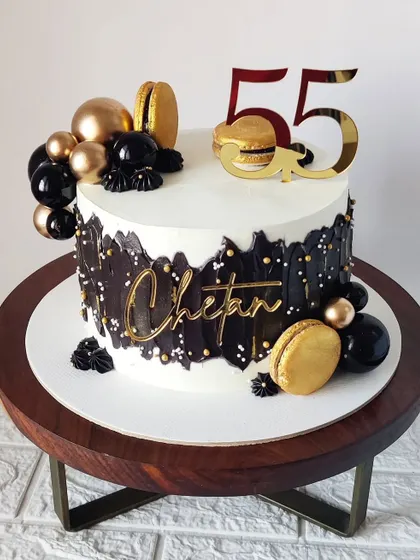 A sophisticated white cake with a black palette knife design and gold accents for a 55th birthday. The addition of golden macarons adds a touch of modern luxury.