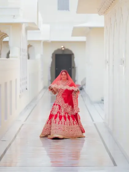 The bride walks down a pristine white corridor in the palace, her red lehenga creating a stunning reflection. This is a powerful, elegant shot that feels like a scene from a movie.