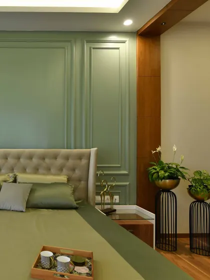 A close-up of a master bedroom featuring a sage green wainscoting accent wall behind a tufted beige headboard. The look is completed with elegant plant stands and rich wooden flooring.
