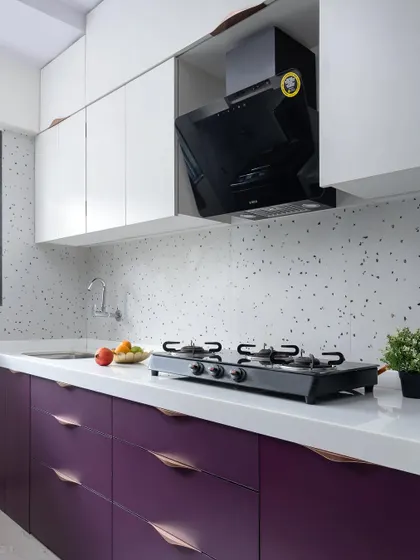 This kitchen from 'The Adobe' project features a bold pop of color with deep purple lower cabinets. The design is balanced with white upper cabinets, a white countertop, and a modern terrazzo-style backsplash.