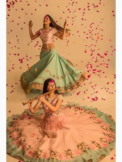 A conceptual fashion shoot for an ethnic wear brand. The composition, with one model playing the flute and another in a joyful pose amidst falling petals, tells a story of grace and celebration, key for a brand lookbook.
