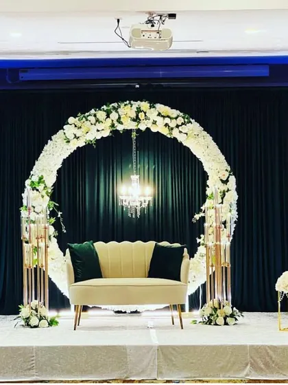 A modern and elegant stage with a circular floral arch. The clean lines, dark backdrop, and minimalist sofa create a sophisticated look for an engagement or reception.