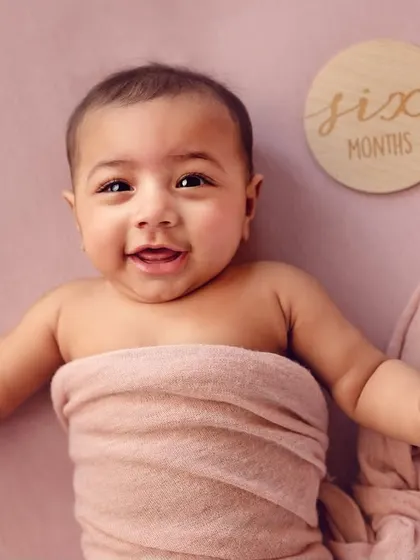 A happy six-month-old baby wrapped in a pink cloth. Milestone sessions for 4 or 6-month-olds are perfect for capturing their developing personalities and adorable expressions.