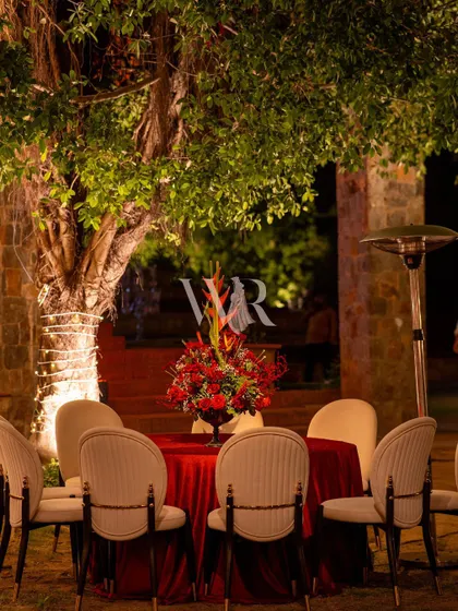 An intimate dining setup for two under a tree. A simple red tablecloth and a centerpiece of red and tropical flowers create a romantic and private corner within the larger celebration.