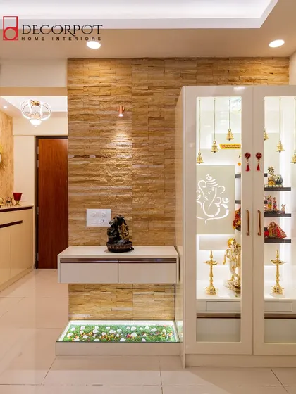 A beautiful pooja unit with glass doors and an etched Ganesha design. The design is set against a stone-clad wall and includes a floating console and a decorative pebble bed below.