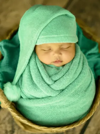 A close-up shot focusing on the baby's peaceful face, snugly wrapped in a green pom-pom hat and nestled in a woven basket.