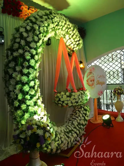 A close-up of a naming ceremony cradle decoration. The crescent moon arch is covered in white chrysanthemums, and a personalized sign with the baby's name adds a special touch.