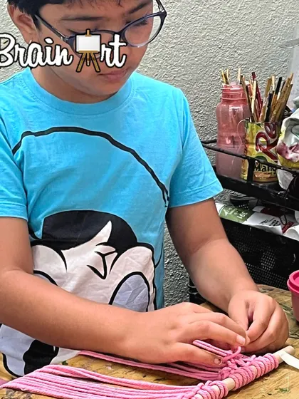 This young artist is carefully weaving pink cord for his macrame project. The concentration on his face shows how engaging and rewarding this craft can be.