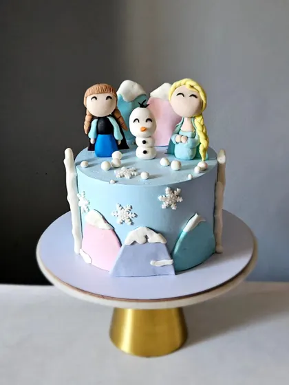 A sweet Frozen-themed cake with handcrafted figurines of Anna, Elsa, and Olaf. The pastel-colored mountains and edible snowflakes create a charming winter wonderland.