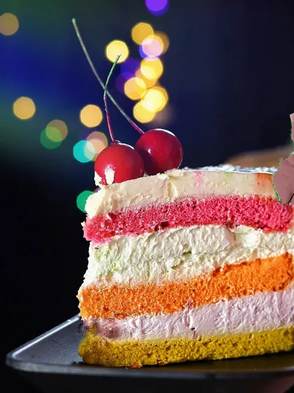 A different perspective of the rainbow pastry, emphasizing the cherry on top and the colorful layers against the dark, festive background.