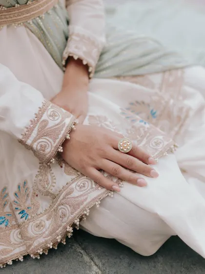 A close up detail shot of Amani's hands, showcasing the delicate embroidery of her outfit and her elegant ring.