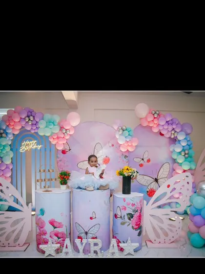 The birthday girl Ayra sits like a princess on her throne at her butterfly-themed first birthday party. The setup features pastel colors and large butterfly wing props.