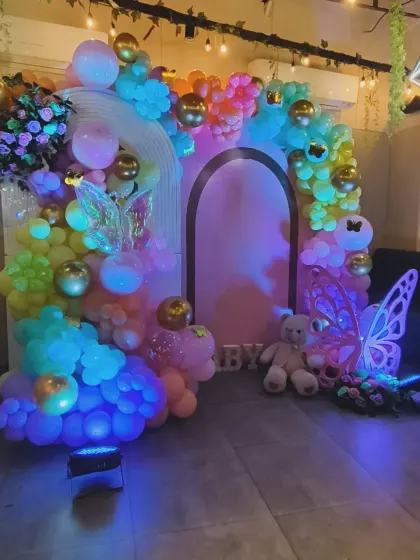 A magical butterfly-themed decoration with a rainbow pastel balloon arch. The setup is enhanced with glowing butterfly wings and a teddy bear, creating a dreamy atmosphere.