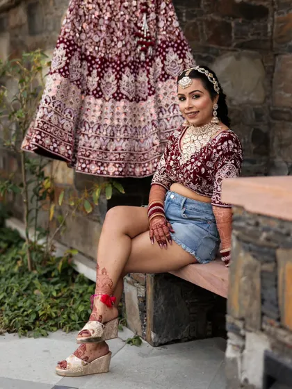 A full-length shot of the bride's modern pre-ceremony look, confidently posing in her blouse and denim shorts next to her hanging lehenga skirt.