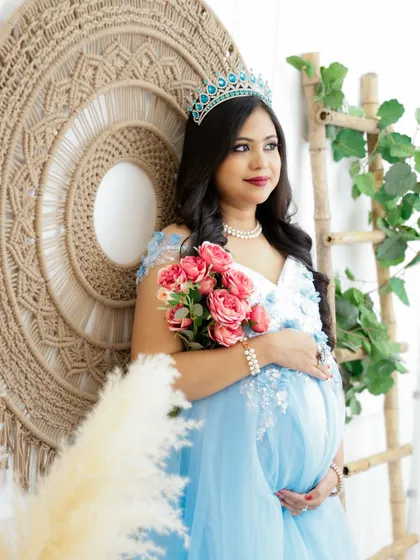 A regal portrait in a boho-chic setting. The mom-to-be wears a tiara and a beautiful blue gown, holding a bouquet of pink roses.