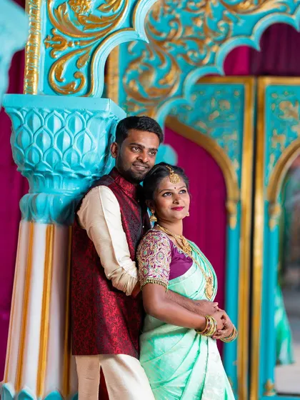 A beautifully dressed couple in traditional Indian wedding attire poses against a vibrant, ornate backdrop, perfect for a reception or sangeet look.