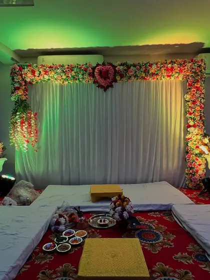 A simple mandap setup for a home or small hall. The backdrop is a white curtain with a floral arch, and the seating is arranged on the floor.
