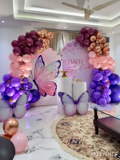 An elegant butterfly theme for a baby shower, set up in a home. This design uses two curved backdrops, large butterfly cutouts, and a rich color palette of purple, pink, and rose gold balloons to create a stunning focal point.