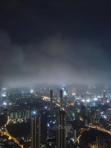 A wide drone shot of Mumbai at night, showing a bank of monsoon clouds rolling over the illuminated cityscape.