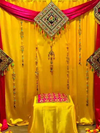 This home Haldi setup uses a combination of yellow and pink drapes with traditional mirror-work hangings. A single seat is prepared for the ceremony, surrounded by marigold flowers on the floor.