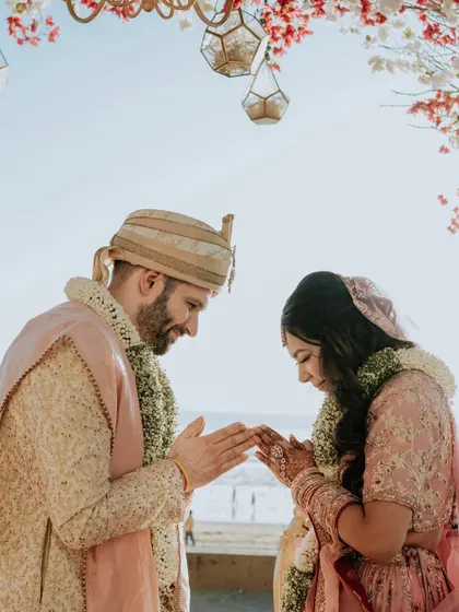 A moment of reverence and connection during a beachside ceremony. The couple's hands are joined in a traditional gesture, their faces close, with the ocean in the background.