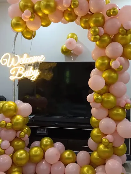 An elegant 'Welcome Baby' balloon arch in pink and gold, designed to frame a TV or fireplace area in a home. The setup is highlighted by a warm white neon sign, making it a beautiful focal point.