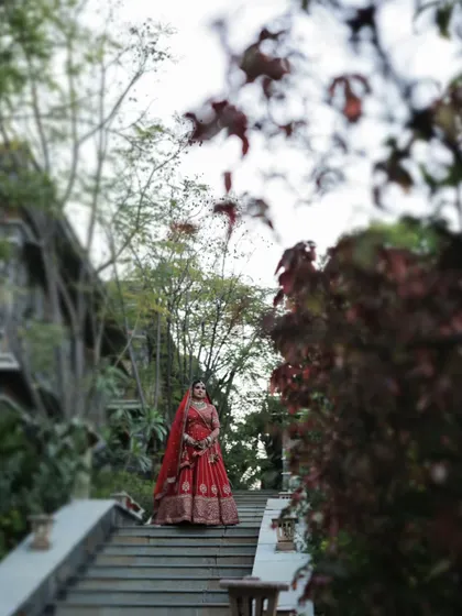 A wide environmental portrait of the bride in her red lehenga, standing on a stone staircase surrounded by lush greenery.