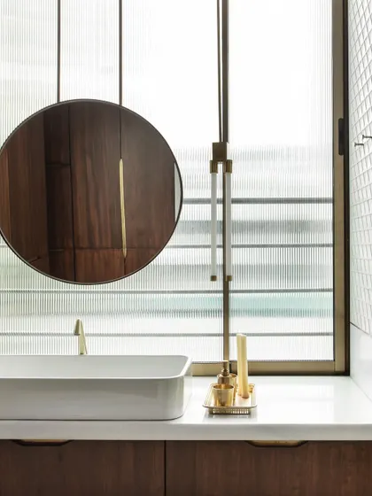 The master washroom features a vessel sink, a bespoke circular mirror, and brass fixtures against a backdrop of fluted glass, balancing privacy with abundant natural light.